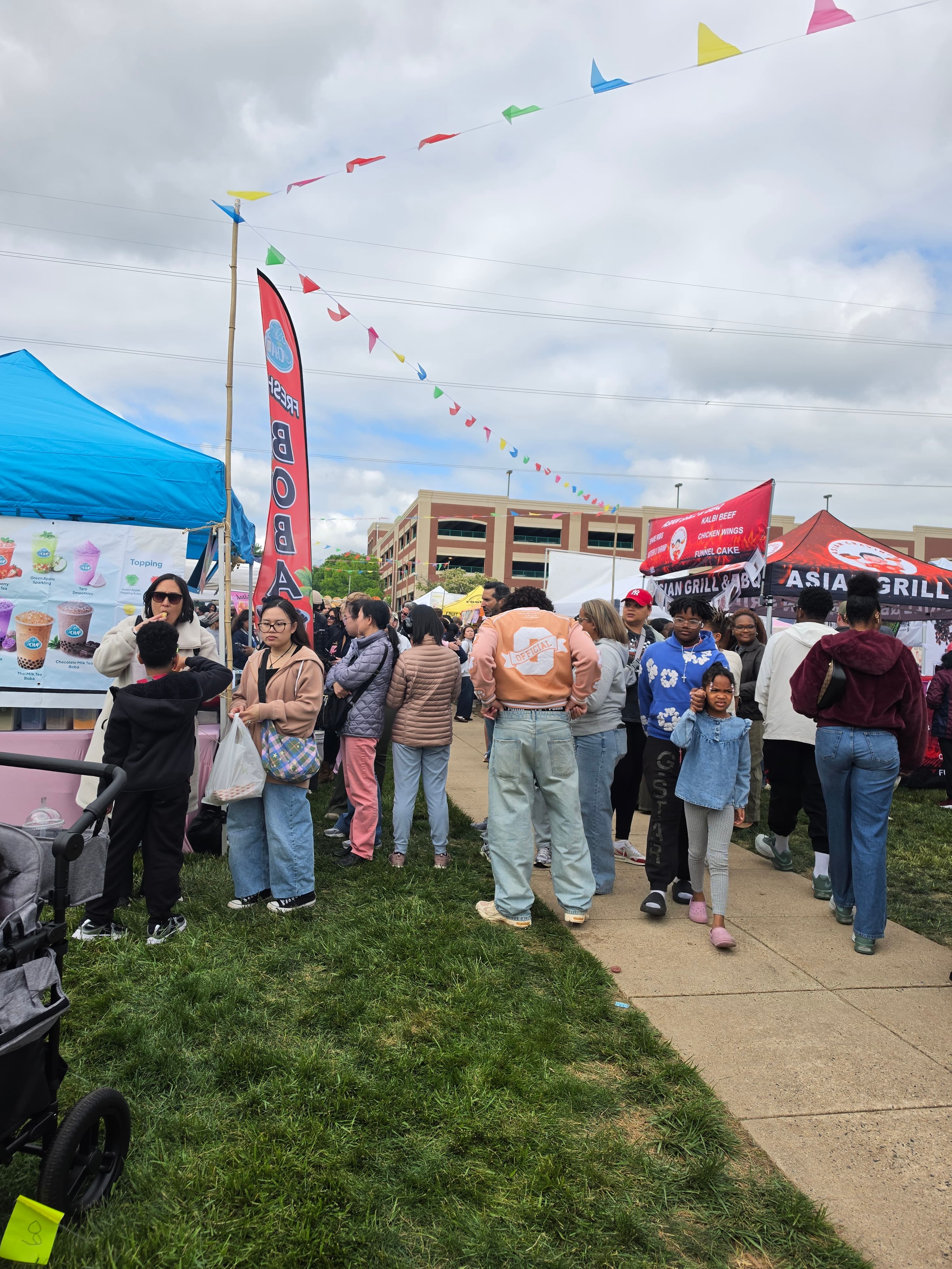 People queue at food stalls under colorful pennant flags at a busy outdoor festival.